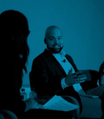 Will Cordery speaks on stage during a plenary session. Ashley Blanchard is sitting next to Will and looking at him smiling.