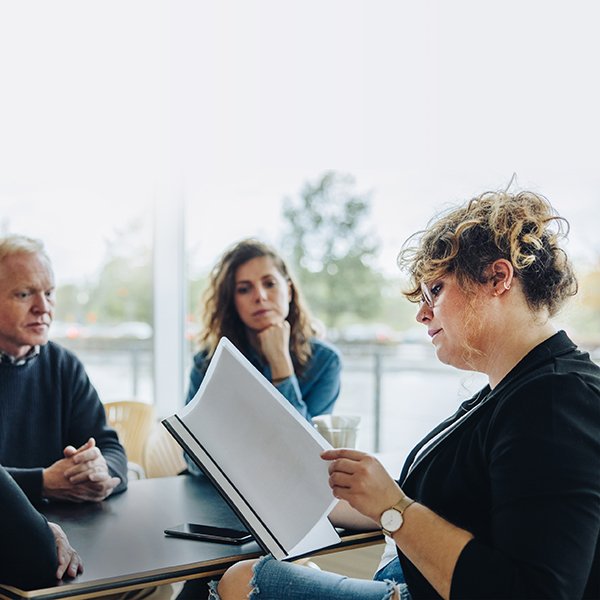 Woman at conference room table reading through paperwork while another man and woman look on.