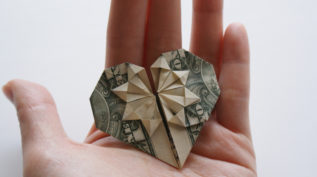 A hand holds a dollar bill that is shaped like an origami heart