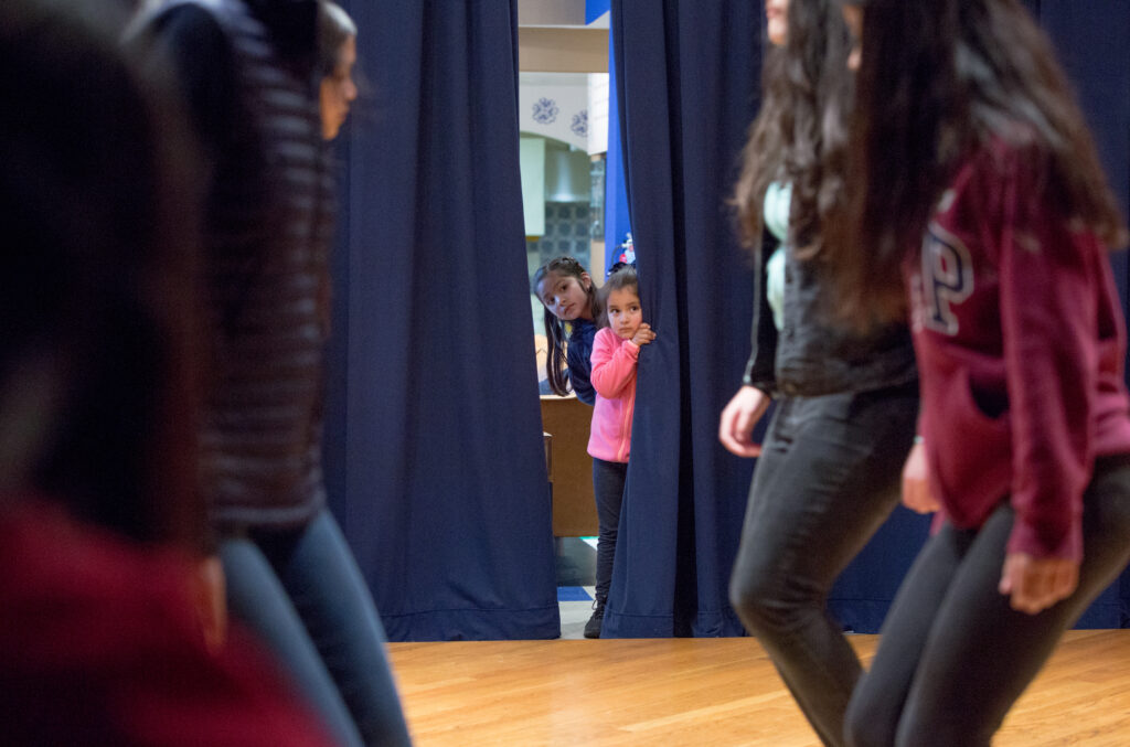 Two young girls peek from behind a curtain into a dance studio where youth are practicing a dance